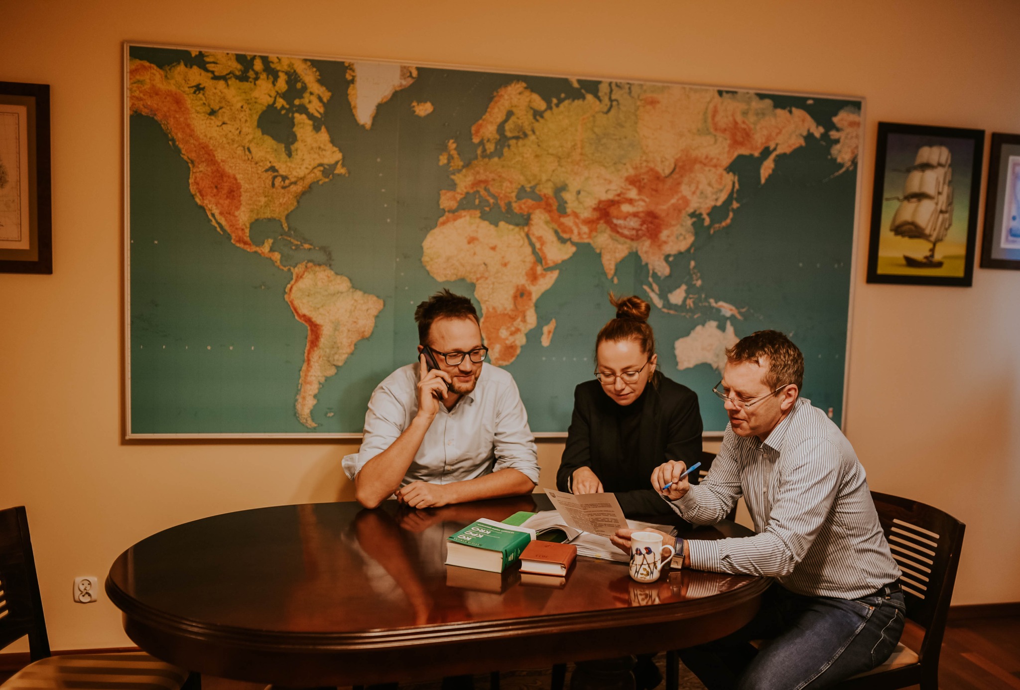 Trzech prawników w okularach omawia dokumenty przy okrągłym drewnianym stole; w tle mapa świata i obrazy.