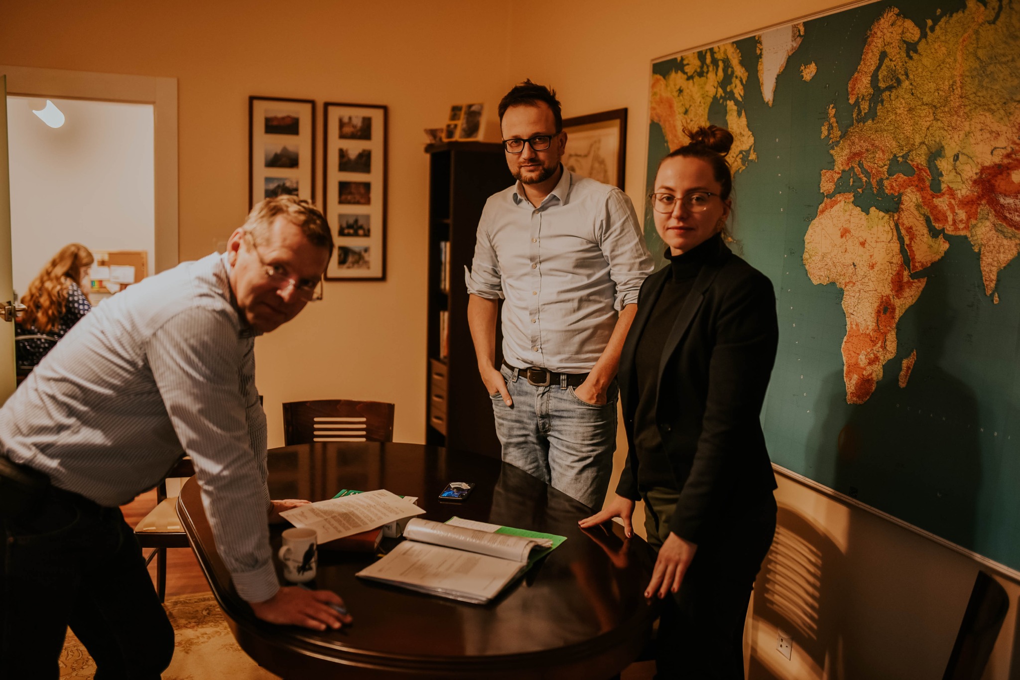 Trzech prawników w biurze omawia dokumenty na okrągłym stole, za nimi mapa świata i zdjęcia na ścianie, jeden z nich trzyma kubek z orłem.