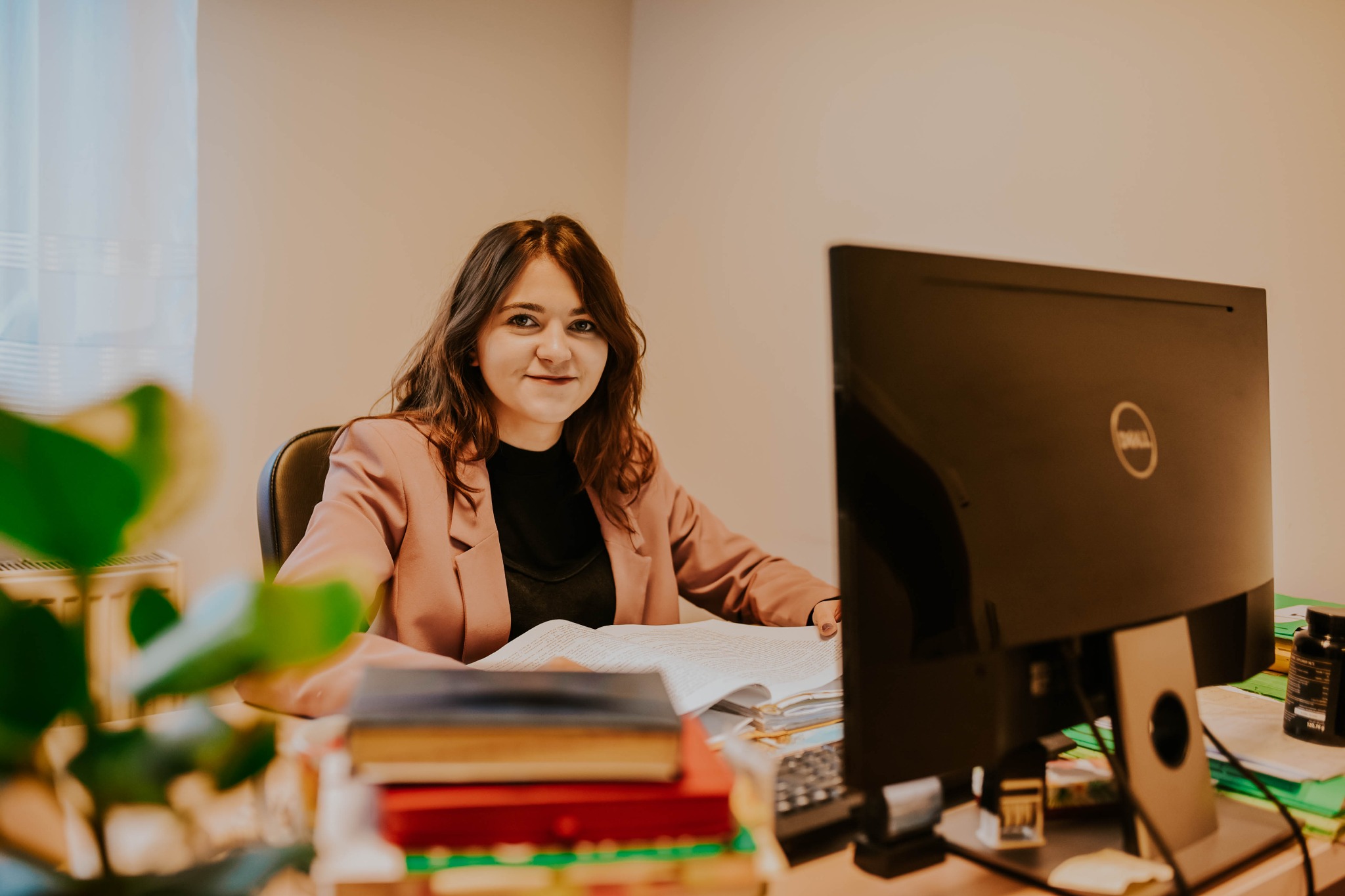 Uśmiechnięta kobieta w różowej marynarce siedzi za biurkiem, przeglądając dokumenty. Na biurku widoczne stosy książek, monitor Dell i roślina doniczkowa.
