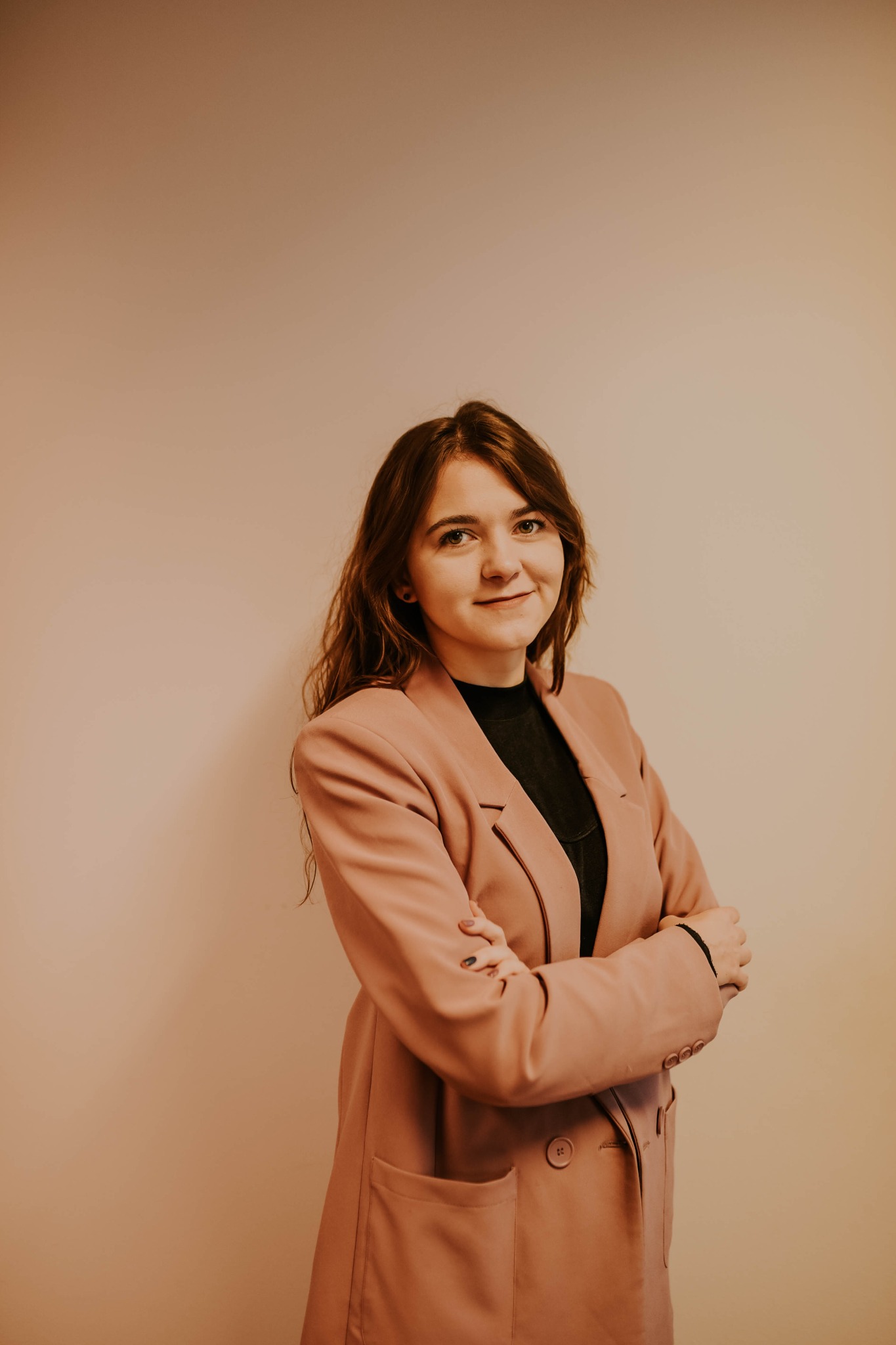 Portret młodej kobiety z długimi, brązowymi włosami, ubranej w różowy, oversize'owy blezer, z rękami skrzyżowanymi na piersiach na tle beżowej ściany. Widoczne subtelne kolczyki w uszach.
