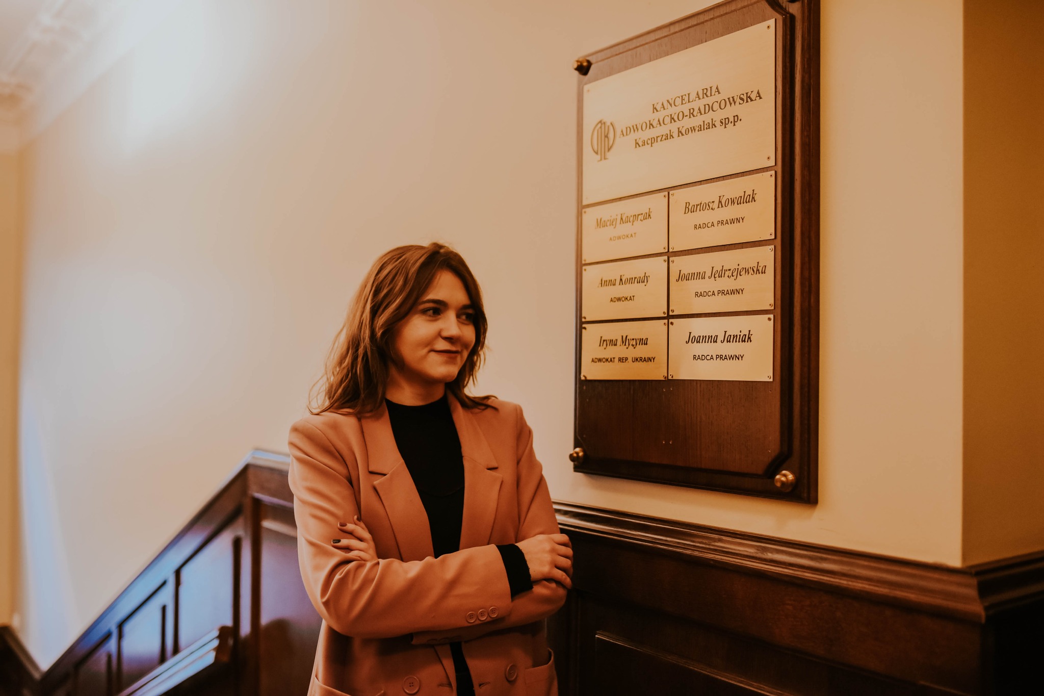 Portret kobiety w różowym żakiecie stojącej na tle tablicy informacyjnej Kancelarii Adwokacko-Radcowskiej z wypisanymi nazwiskami adwokatów i radców prawnych.