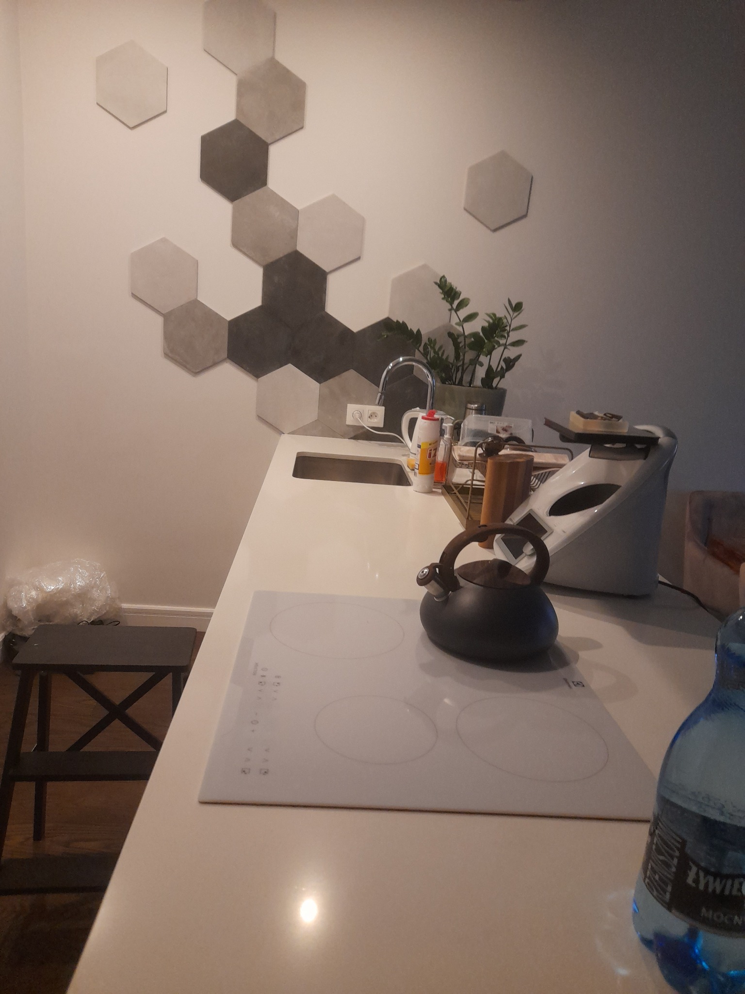 Nowoczesna kuchnia z białym blatem, czajnikiem na płycie indukcyjnej, ozdobnymi heksagonalnymi płytkami na ścianie i rośliną doniczkową w tle.