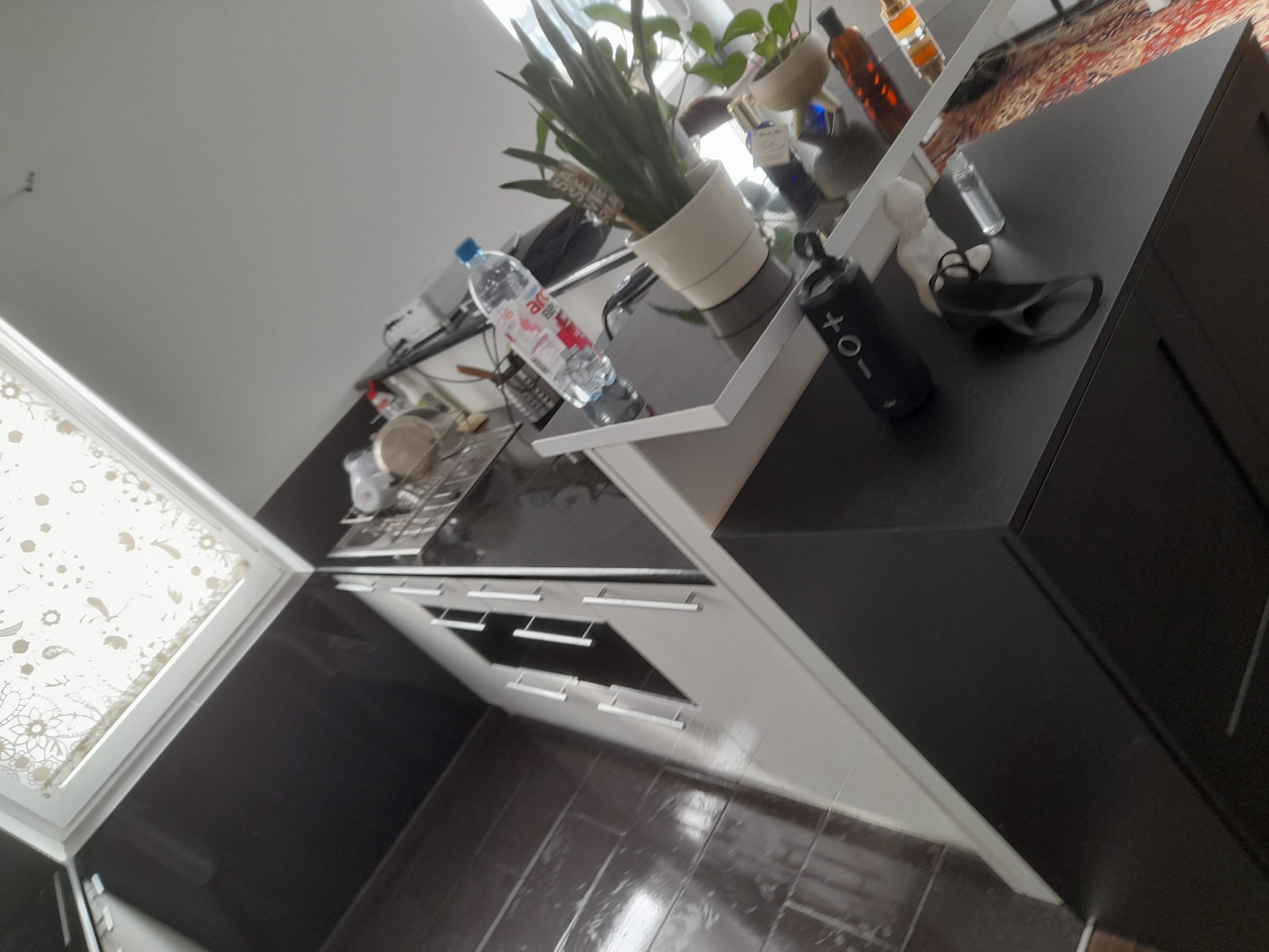 Ukośne ujęcie nowoczesnej kuchni z ciemnymi szafkami i jasnymi blatami, na których stoją roślina doniczkowa, butelka wody i głośnik. Widoczny fragment okna z wzorzystą zasłoną.