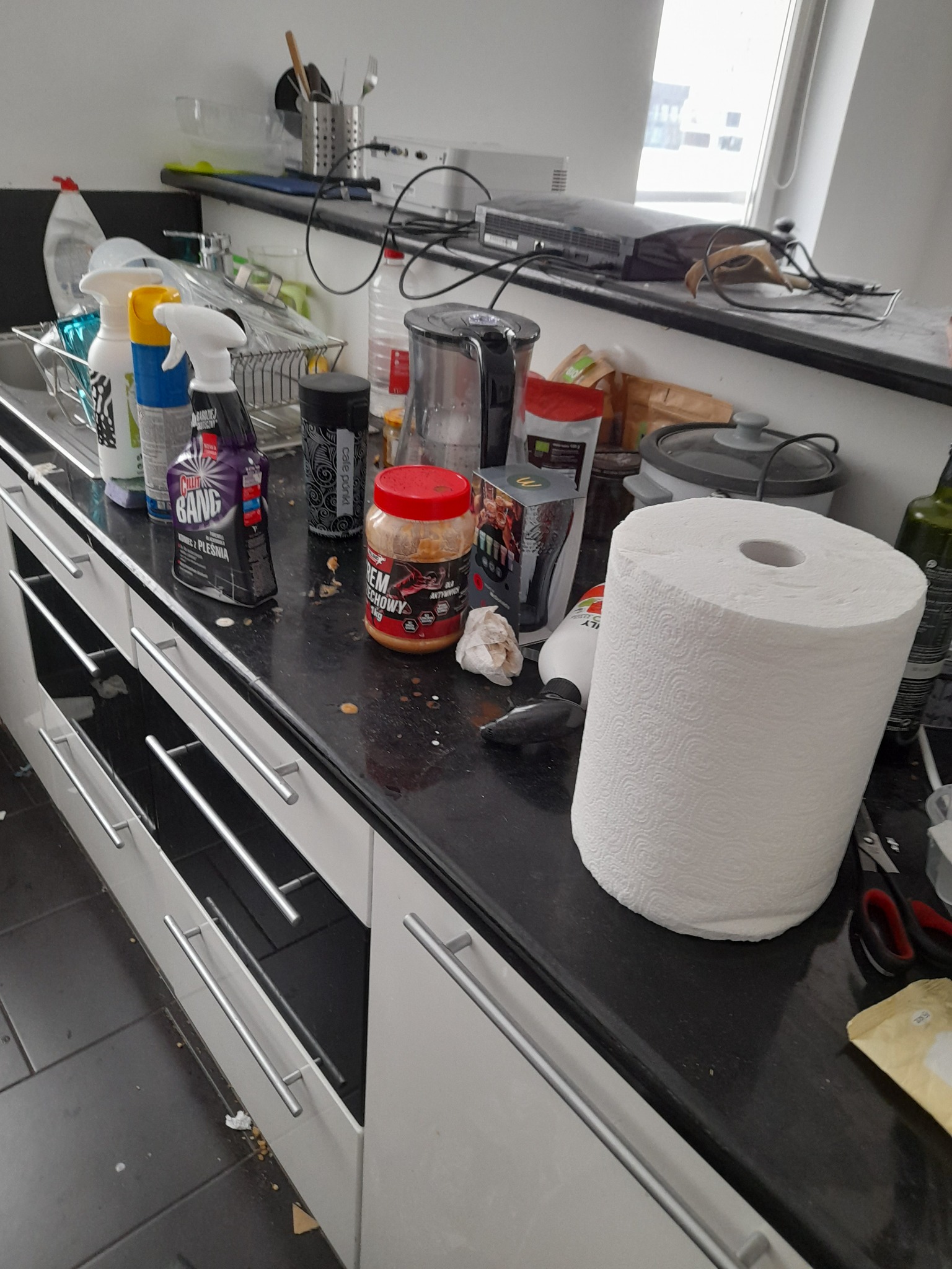 Zabrudzony blat kuchenny z widocznymi plamami, resztkami jedzenia i butelkami środków czyszczących, rolka ręcznika papierowego i inne przedmioty codziennego użytku. Widok na okno w tle.