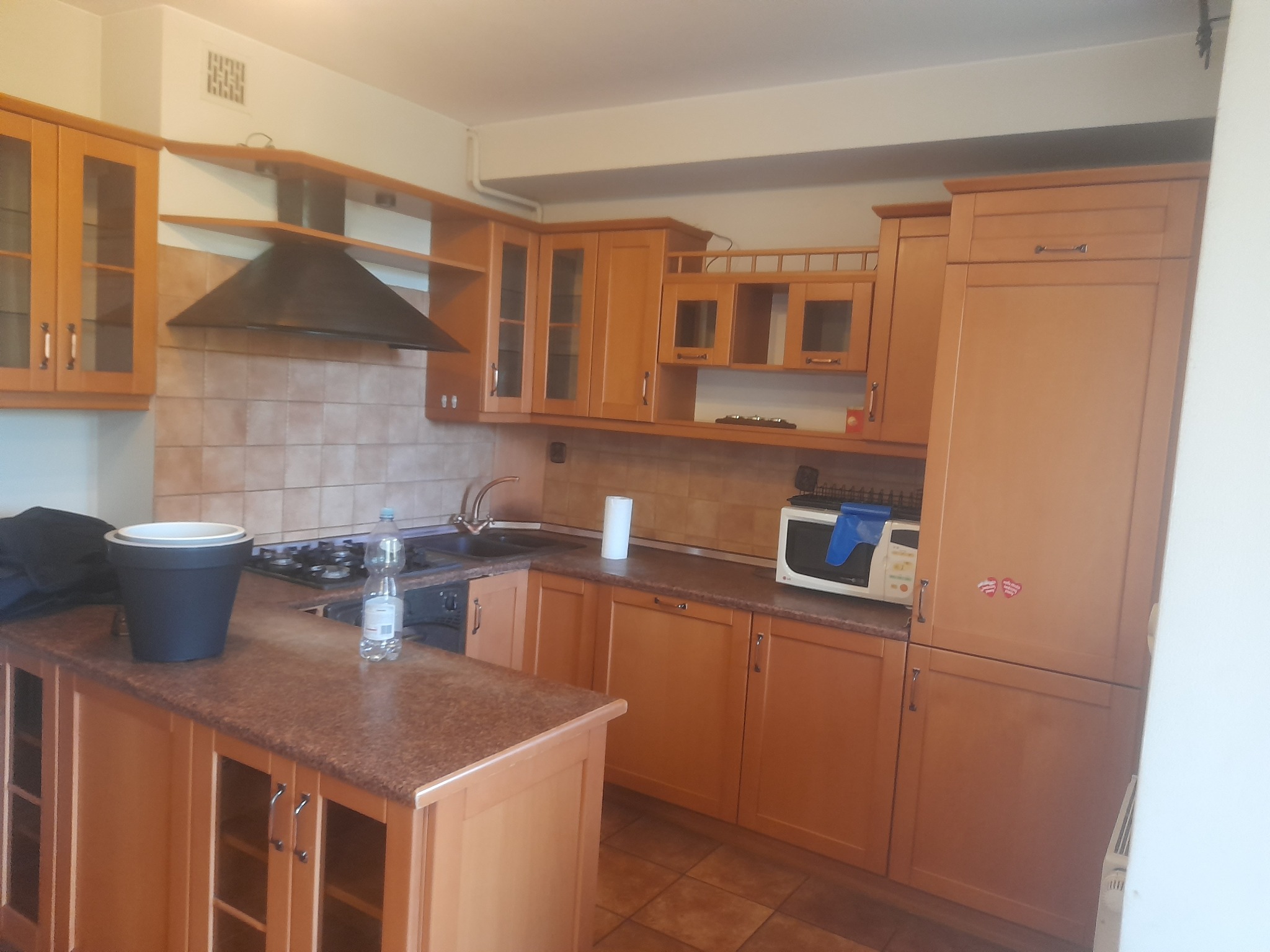 Kuchnia z drewnianymi szafkami przed sprzątaniem, widoczny czarny worek na śmieci i butelka wody na blacie.