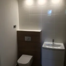 Nowoczesna łazienka z białymi płytkami, podwieszaną toaletą i umywalką z szafką, oświetlona halogenami, z brązową zabudową za toaletą i brązowymi płytkami na podłodze.