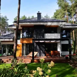 Dwukondygnacyjny dom murowany z płaskim dachem i elementami drewnianymi na elewacji, otoczony lasem sosnowym, widok z perspektywy ogrodu z kwitnącymi hortensjami na pierwszym planie.