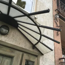 Metalowa konstrukcja z przezroczystym zadaszeniem nad wejściem do budynku, widoczna lampa ścienna i numer budynku.