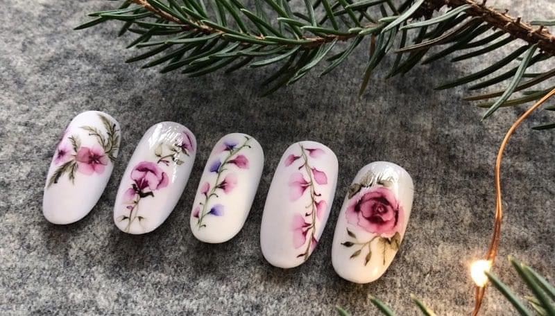 Pięć białych tipsów z ręcznie malowanymi motywami kwiatowymi w odcieniach różu i fioletu, ułożonych na szarym tle z gałązką świerku i delikatnym oświetleniem.