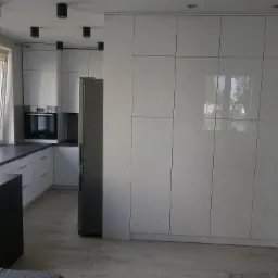 Jasna kuchnia na wymiar z białymi, lakierowanymi frontami, szarym blatem i zabudowaną szafą wnękową z połyskiem, widoczna drabina malarska.