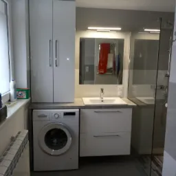 Funkcjonalna aranżacja łazienki w Elblągu: biała szafa wisząca nad pralką, umywalka z szafką i kabina prysznicowa z widocznymi kafelkami.