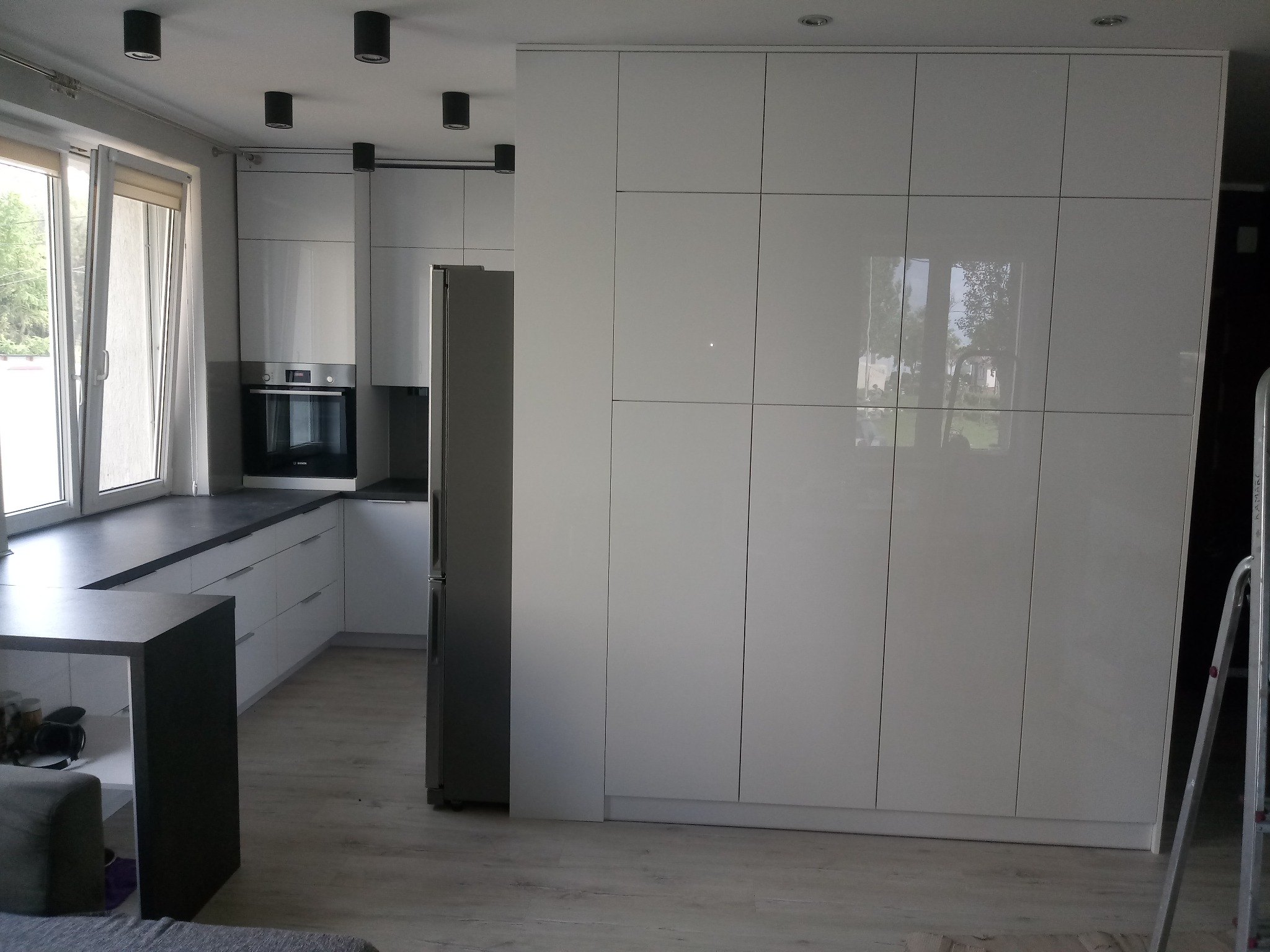 Jasna kuchnia na wymiar z białymi, lakierowanymi frontami, szarym blatem i zabudowaną szafą wnękową z połyskiem, widoczna drabina malarska.