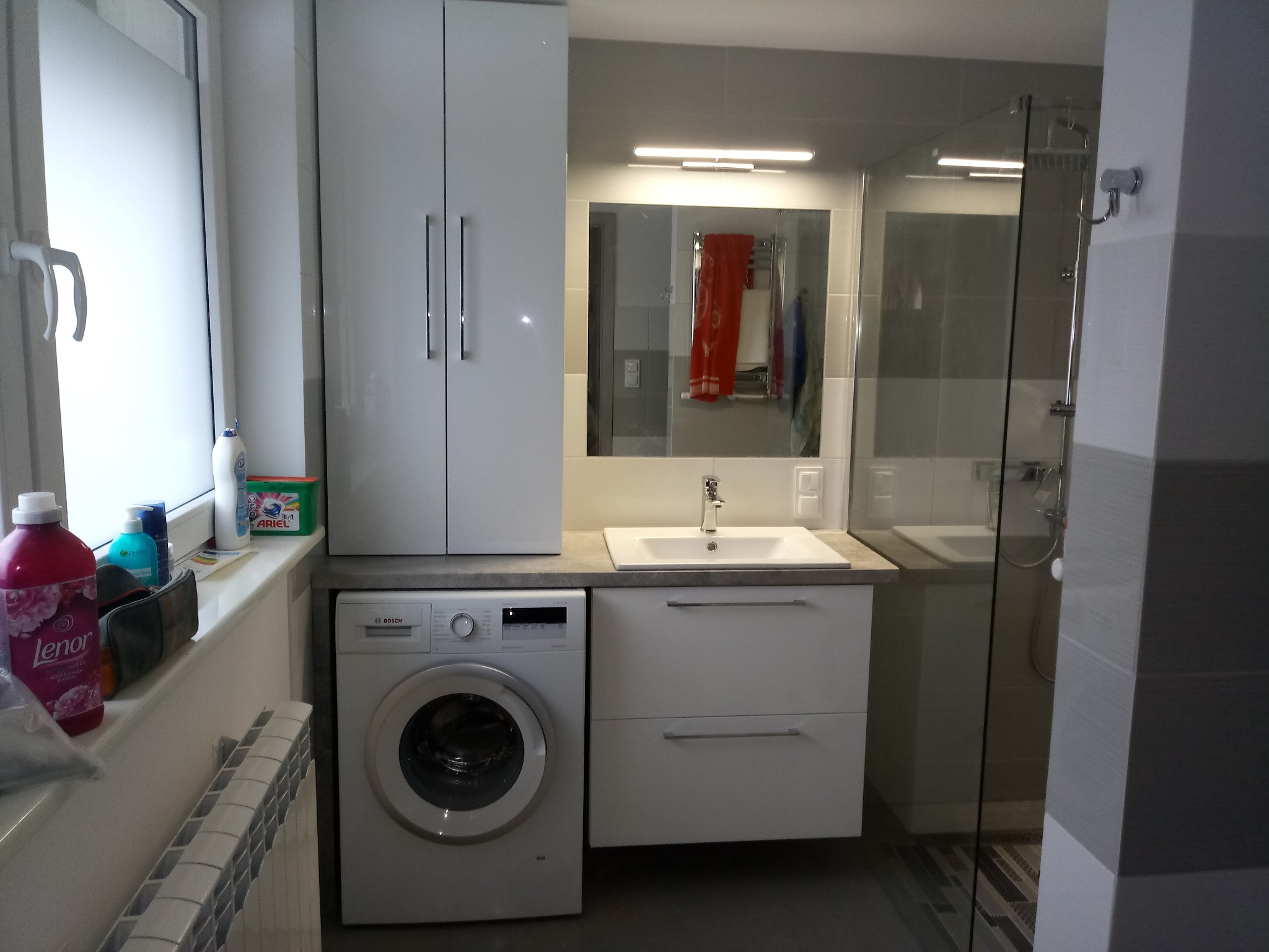 Funkcjonalna aranżacja łazienki w Elblągu: biała szafa wisząca nad pralką, umywalka z szafką i kabina prysznicowa z widocznymi kafelkami.