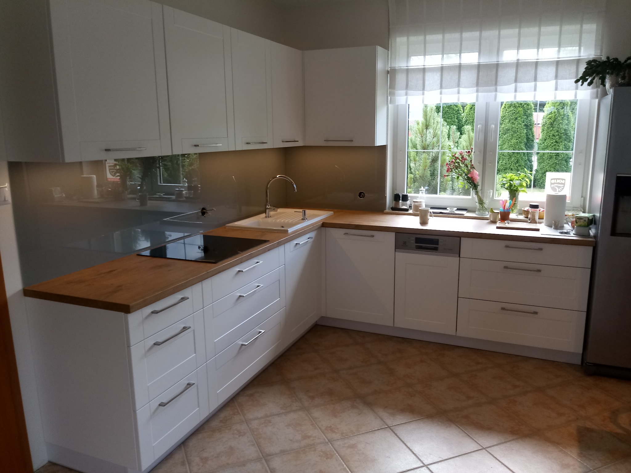 Jasna kuchnia z białymi szafkami i drewnianymi blatami, widok na okno z kwiatami i zielenią, płytki podłogowe w odcieniach brązu.