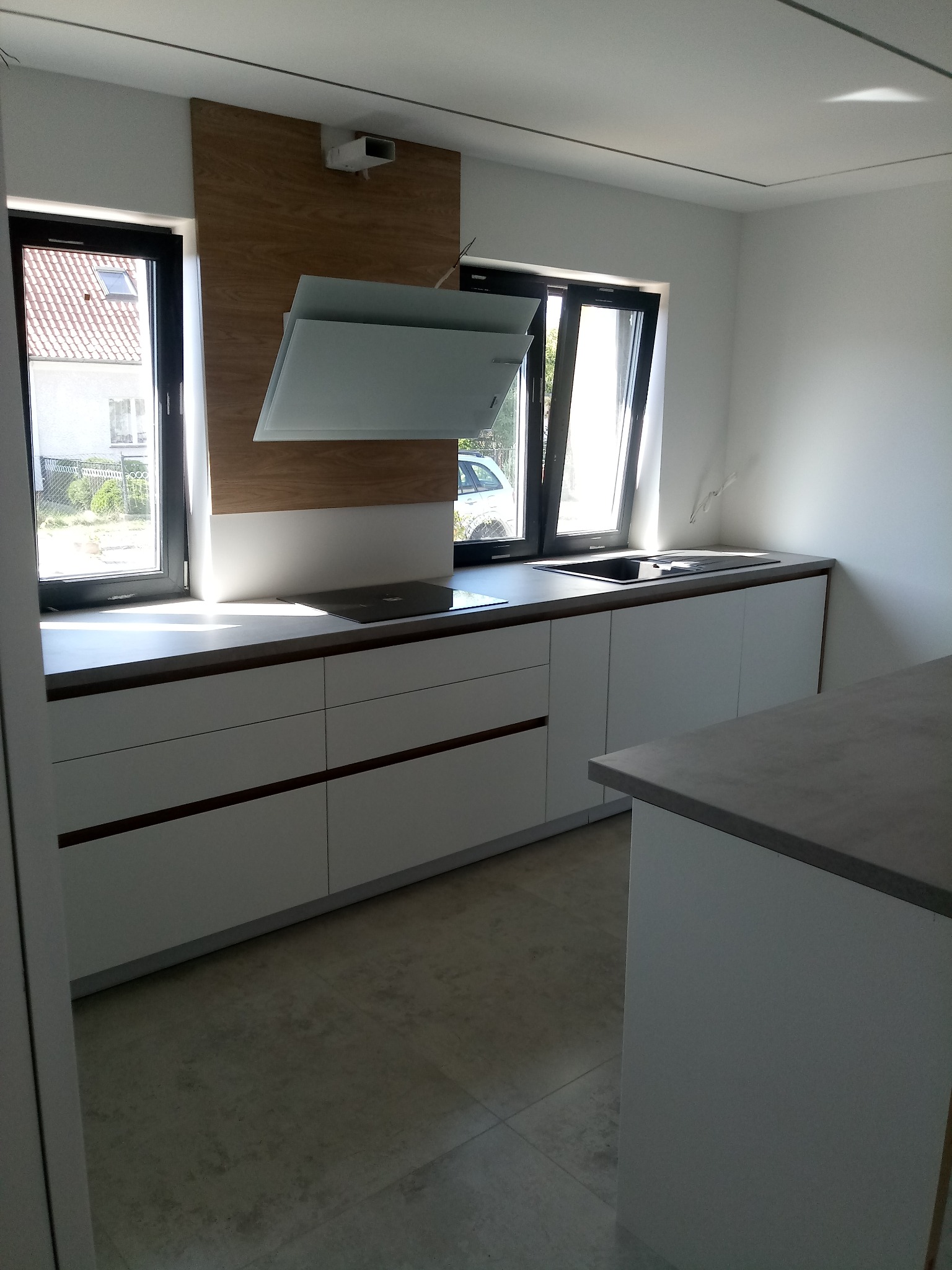 Nowoczesna, jasna kuchnia z białymi szafkami i drewnianym panelem za szklanym okapem. Blat w kolorze szarości. Dwa okna w czarnych ramach wpuszczają dużo światła.