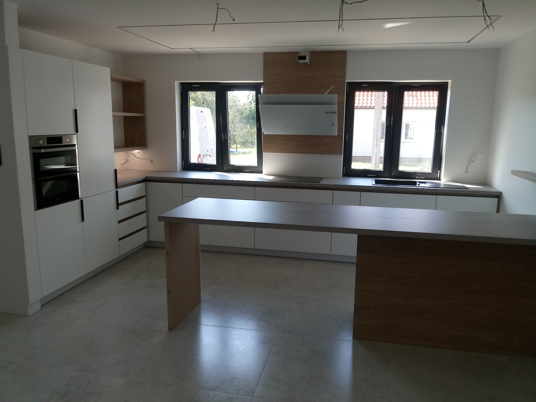 Nowoczesna kuchnia z białymi szafkami i drewnianym blatem wyspowym. Dwa okna w tle wpuszczają naturalne światło. Minimalistyczny design z elementami drewna.