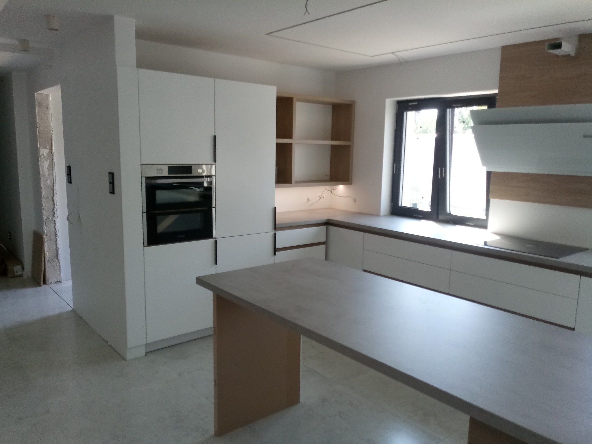 Jasna kuchnia z białymi szafkami i drewnianymi akcentami, wyspa kuchenna z blatem w kolorze betonu, nowoczesny design, oświetlenie LED pod szafkami.