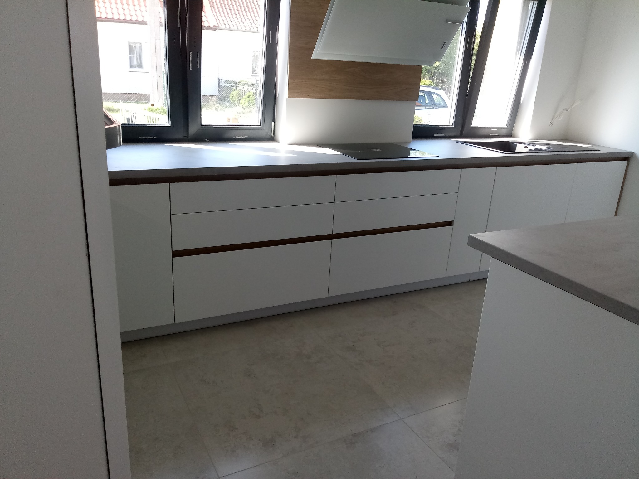 Nowoczesna, minimalistyczna kuchnia z białymi frontami i drewnianymi akcentami. Blat w odcieniach szarości, okno z czarną ramą. Widoczny fragment płyty indukcyjnej i zlewu.