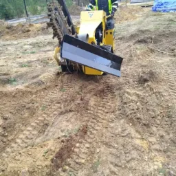 Operator w odblaskowej kamizelce obsługuje żółtą mini-koparkę łańcuchową Vermeer na nierównym, błotnistym terenie budowy w pobliżu lasu i budynku w stanie surowym.