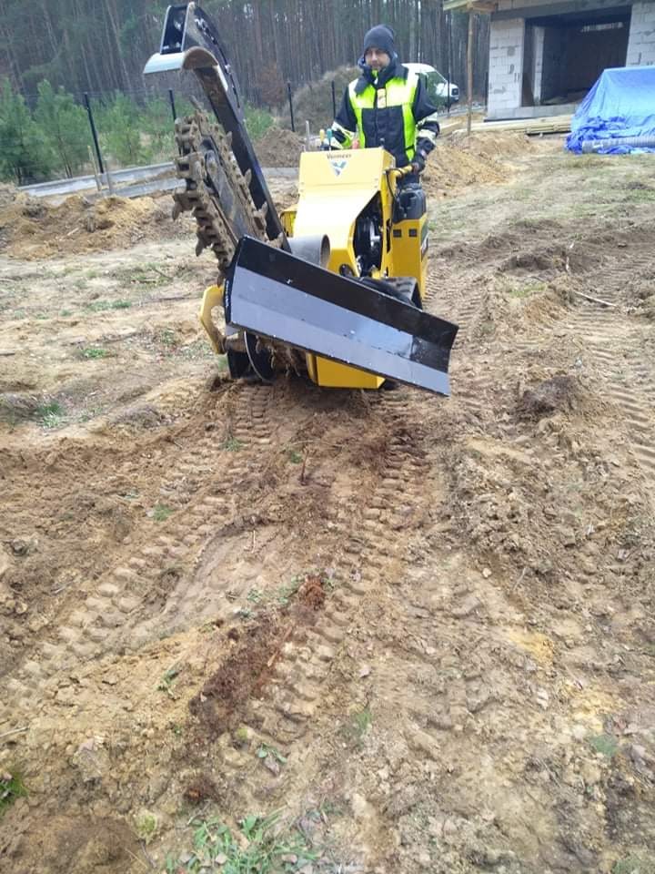 Operator w odblaskowej kamizelce obsługuje żółtą mini-koparkę łańcuchową Vermeer na nierównym, błotnistym terenie budowy w pobliżu lasu i budynku w stanie surowym.