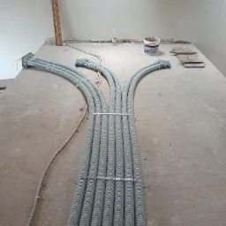 Spięte rury karbowane w szarej osłonie, rozchodzące się w trzech kierunkach na surowym betonowym podłożu w trakcie prac instalacyjnych, widoczne elementy mocujące i puszki przyłączeniowe.