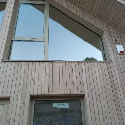 Elewacja budynku z jasnego drewna z zamontowanymi oknami o nietypowych kształtach, widoczny alarm zewnętrzny z kamerą.