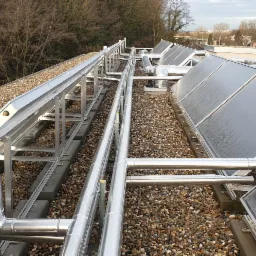 Instalacja solarna na dachu pokrytym drobnymi kamieniami, widoczne rury i panele słoneczne w metalowych ramach, drzewa w tle.