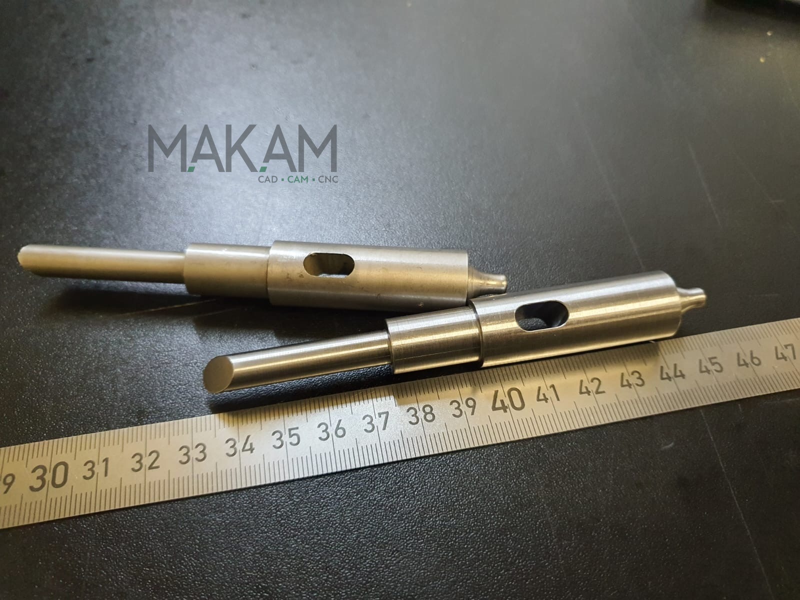 Dwa metalowe trzpienie z otworem, leżące obok miarki. Widoczne logo firmy MAK AM CAD CAM CNC. Precyzyjna obróbka metali, serwis narzędzi.