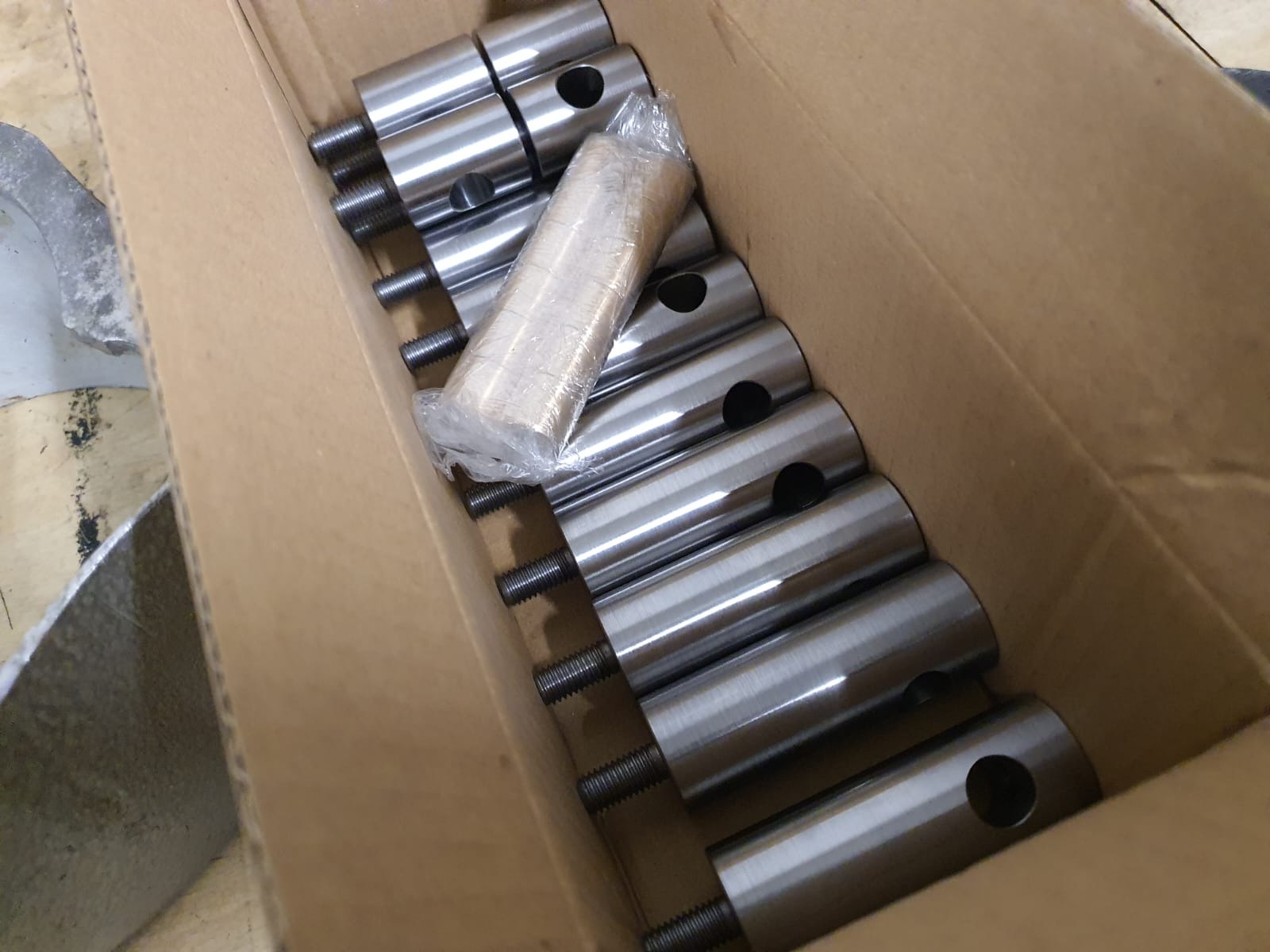 Metalowe, cylindryczne elementy z gwintem i otworem, ułożone w kartonowym pudełku. Część z nich zapakowana w folię. Elementy prawdopodobnie do elektronarzędzi.