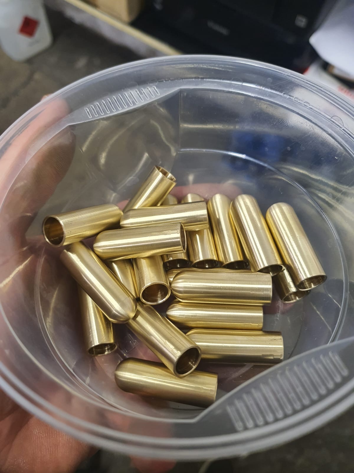 Mosiężne tulejki w przezroczystym pojemniku, prawdopodobnie części zamienne do elektronarzędzi, widoczne ślady użytkowania na pojemniku.