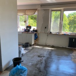 Robert Komenda - Wnętrze w trakcie wylewania posadzki samopoziomującej, widoczne worki z zaprawą, narzędzia i wiadro, okna z widokiem na zieleń