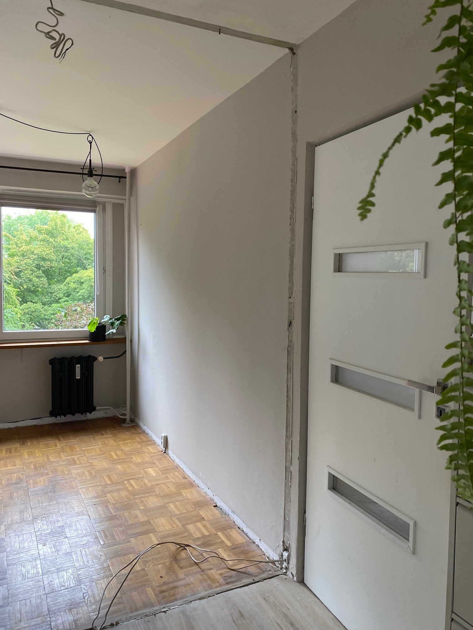 Pokój w trakcie remontu: ściany przygotowane do malowania, widoczne ślady po szpachlowaniu i gładziach, drewniana podłoga parkietowa, białe drzwi z przeszkleniami i wiszący przewód elektryczny...