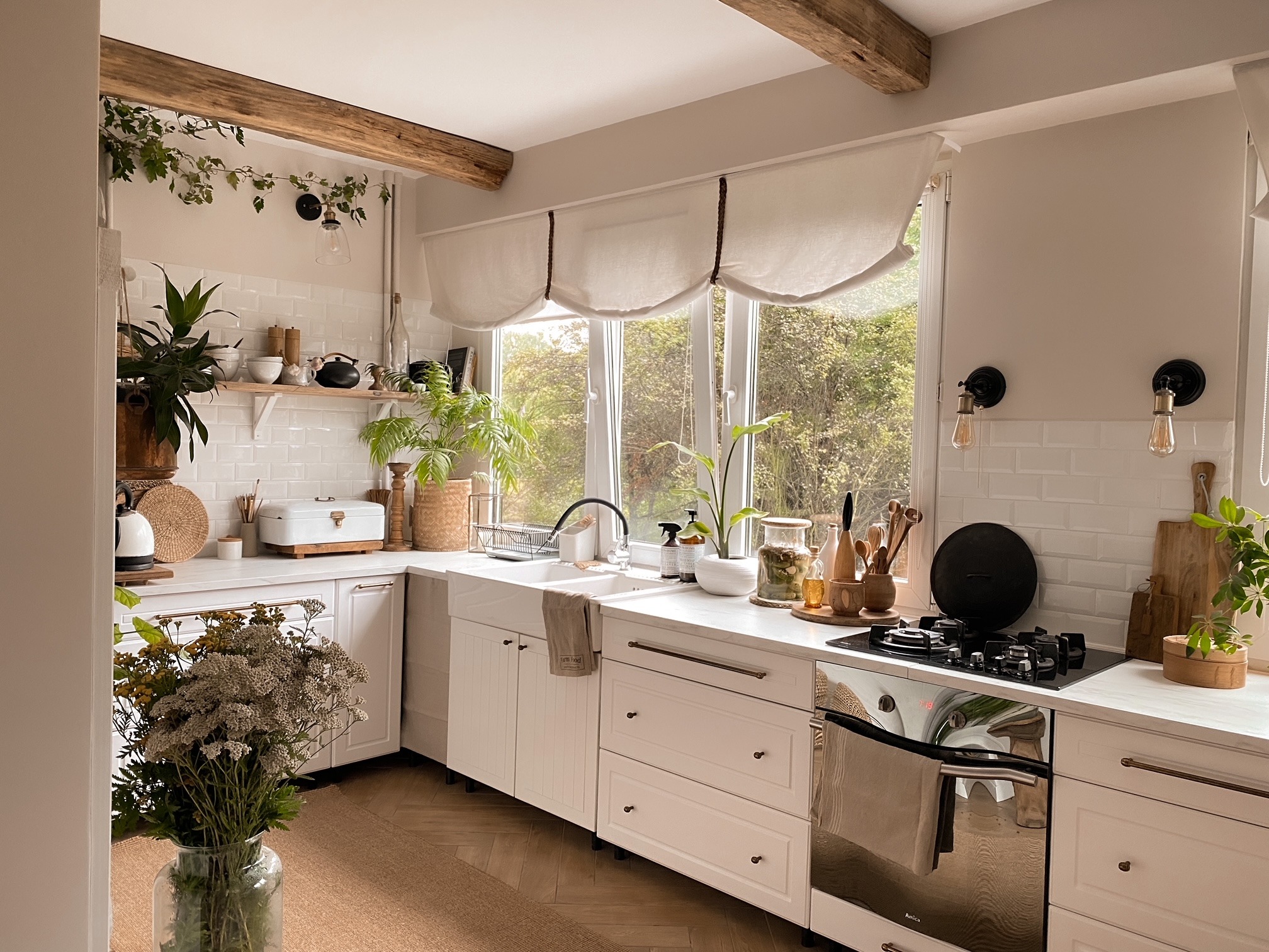 Jasna kuchnia w stylu rustykalnym z drewnianymi belkami stropowymi, białymi szafkami, ceramicznym zlewem farmerskim i licznymi roślinami doniczkowymi oraz bukietem kwiatów w szklanym wazonie...