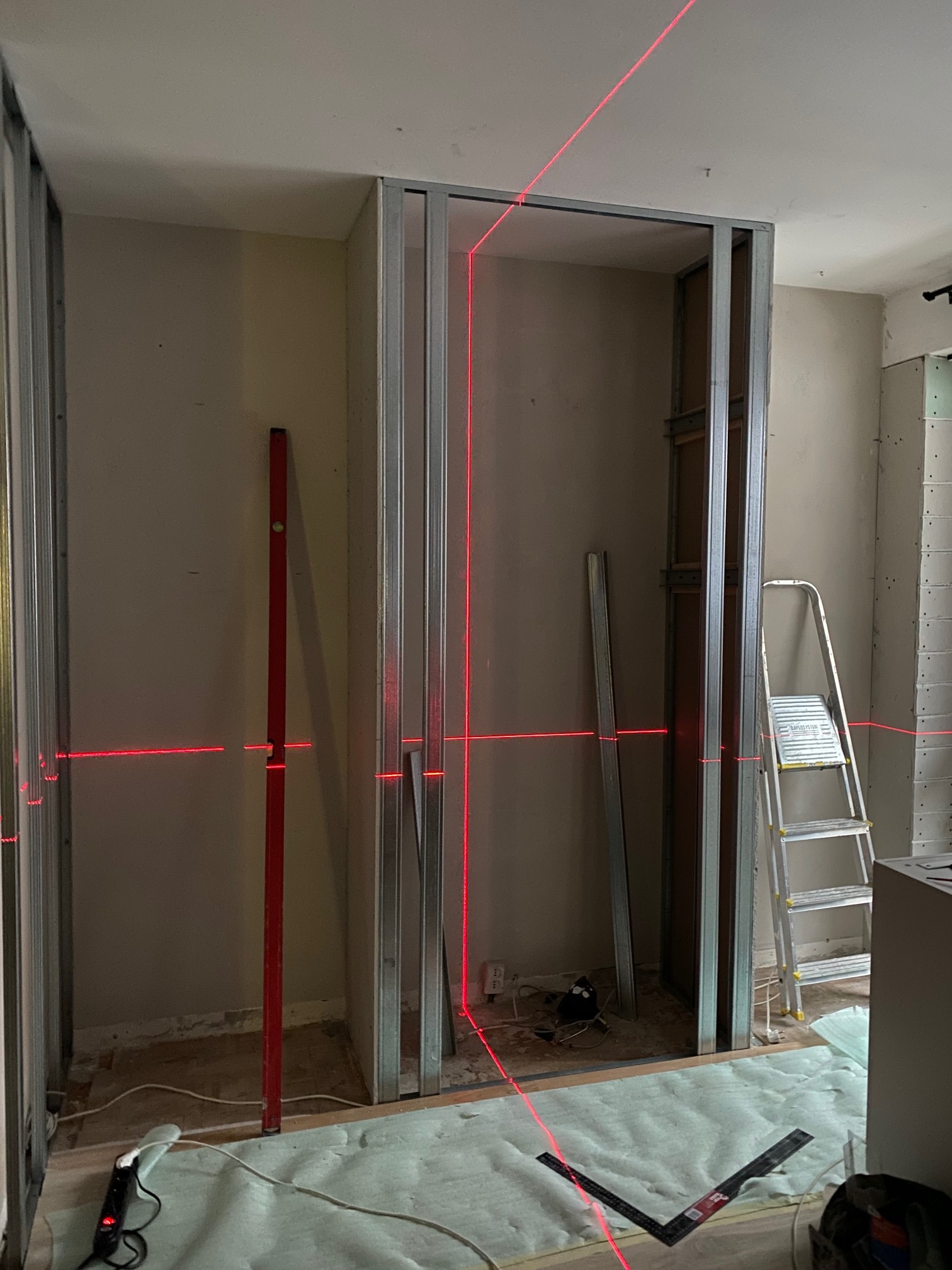 Montaż metalowej konstrukcji ścianki działowej z użyciem poziomicy laserowej, widoczne profile, poziomica pionowa, drabina i narzędzia na podłodze zabezpieczonej folią.