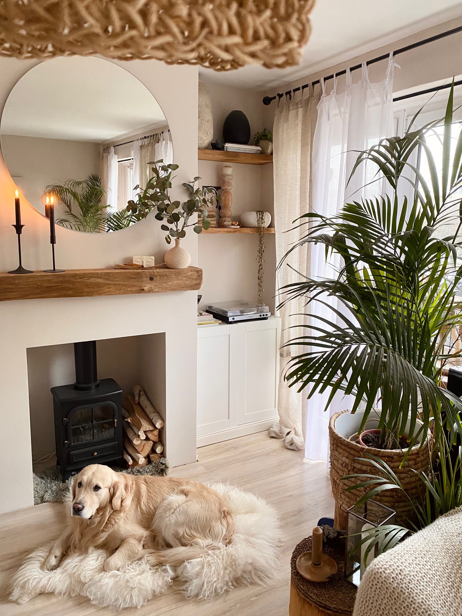 Jasne wnętrze salonu z kominkiem, drewnianą belką, okrągłym lustrem i psem Golden Retriever leżącym na futrzanym dywaniku, z roślinami w tle.
