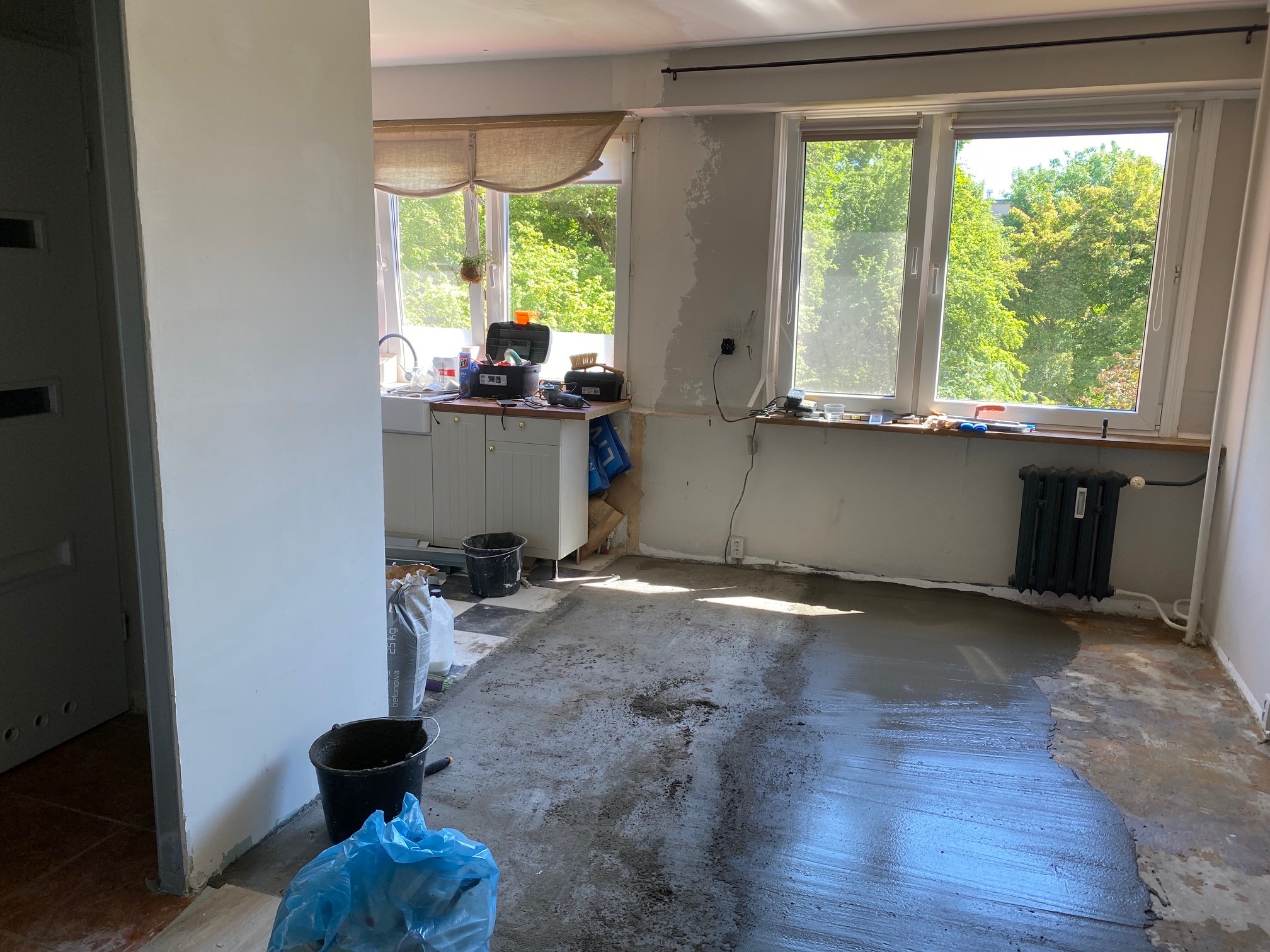 Wnętrze w trakcie wylewania posadzki samopoziomującej, widoczne worki z zaprawą, narzędzia i wiadro, okna z widokiem na zieleń