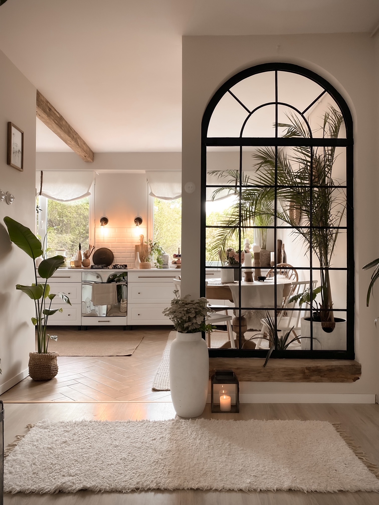 Jasne wnętrze domu z widokiem na kuchnię przez dekoracyjne okno w stylu loft, z roślinami doniczkowymi i dywanem typu shaggy na podłodze.