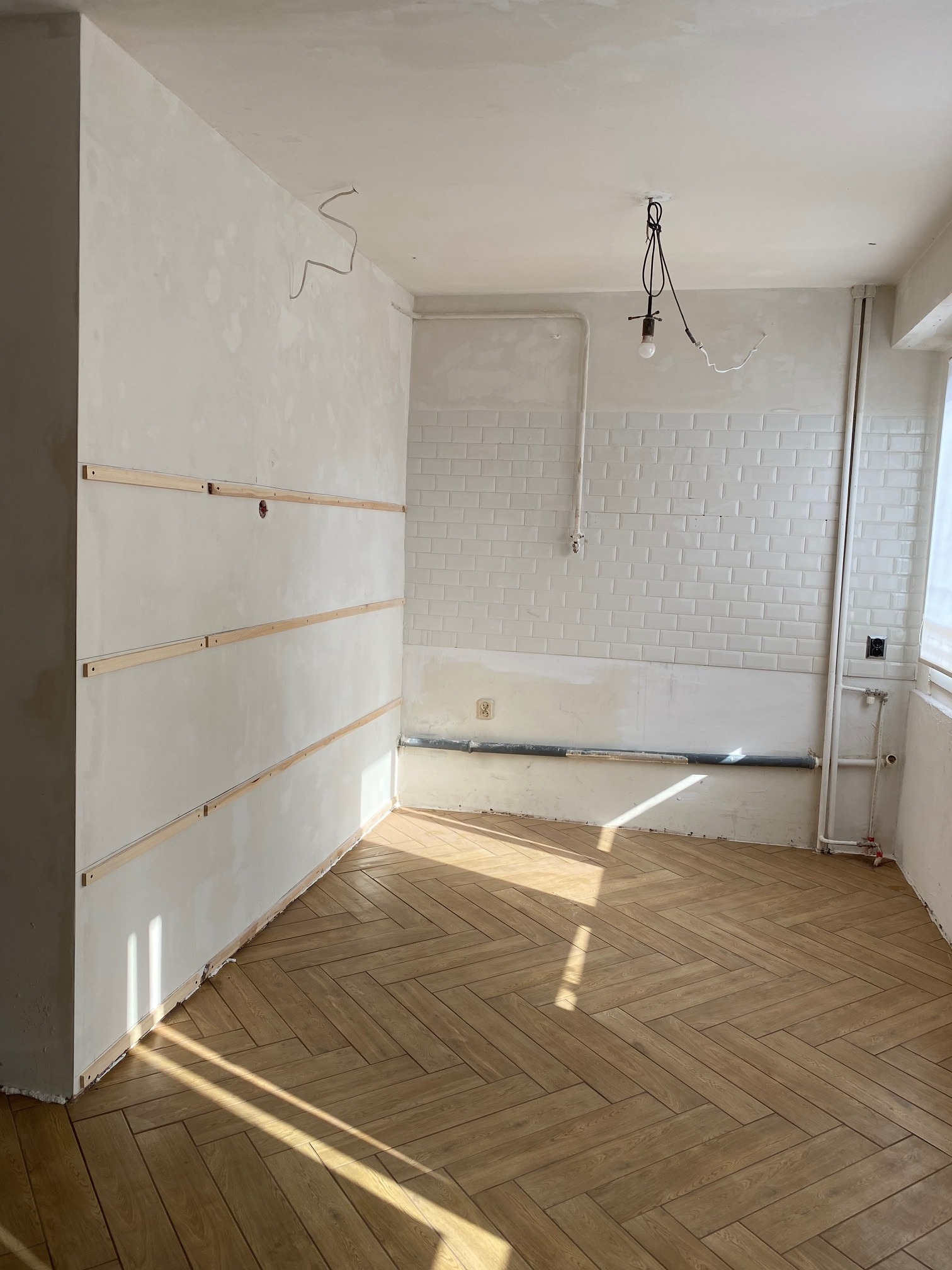 Pokój w trakcie prac wykończeniowych: ściany przygotowane pod malowanie z listwami poziomymi, parkiet ułożony w jodełkę, widoczne instalacje elektryczne i rury.