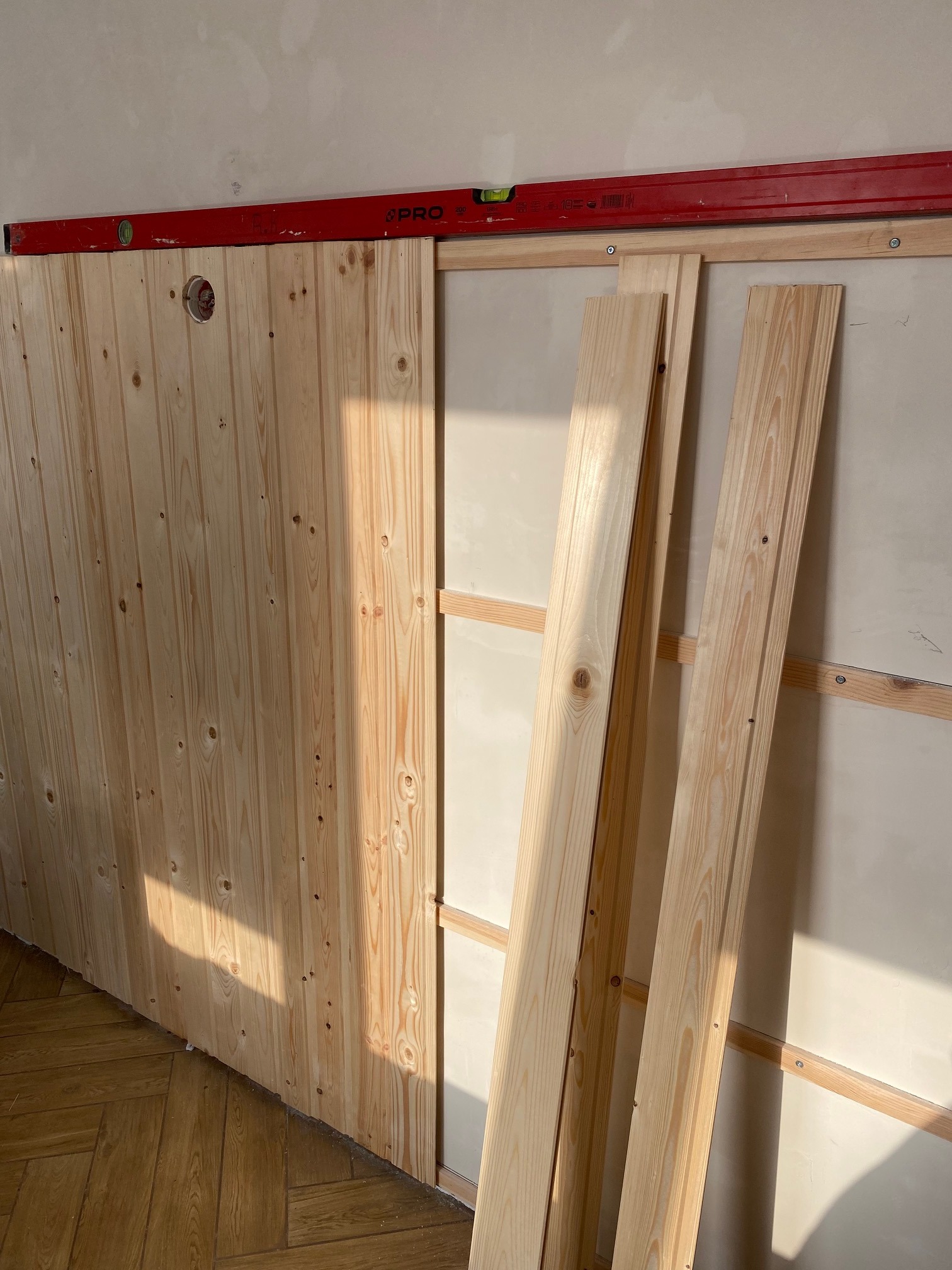 Montaż boazerii z jasnego drewna na ścianie, widoczna poziomica i listwy na podłodze, światło słoneczne pada na drewno.