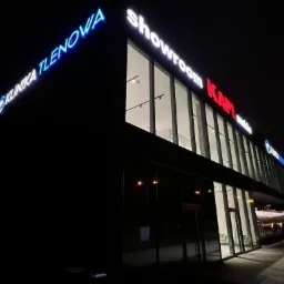 Oświetlony budynek nocą z neonowymi szyldami 'KLINIKA TLENOWA' (niebieski) i 'showroom KAPI meble' (czerwony i biały), nowoczesna architektura z dużymi oknami.