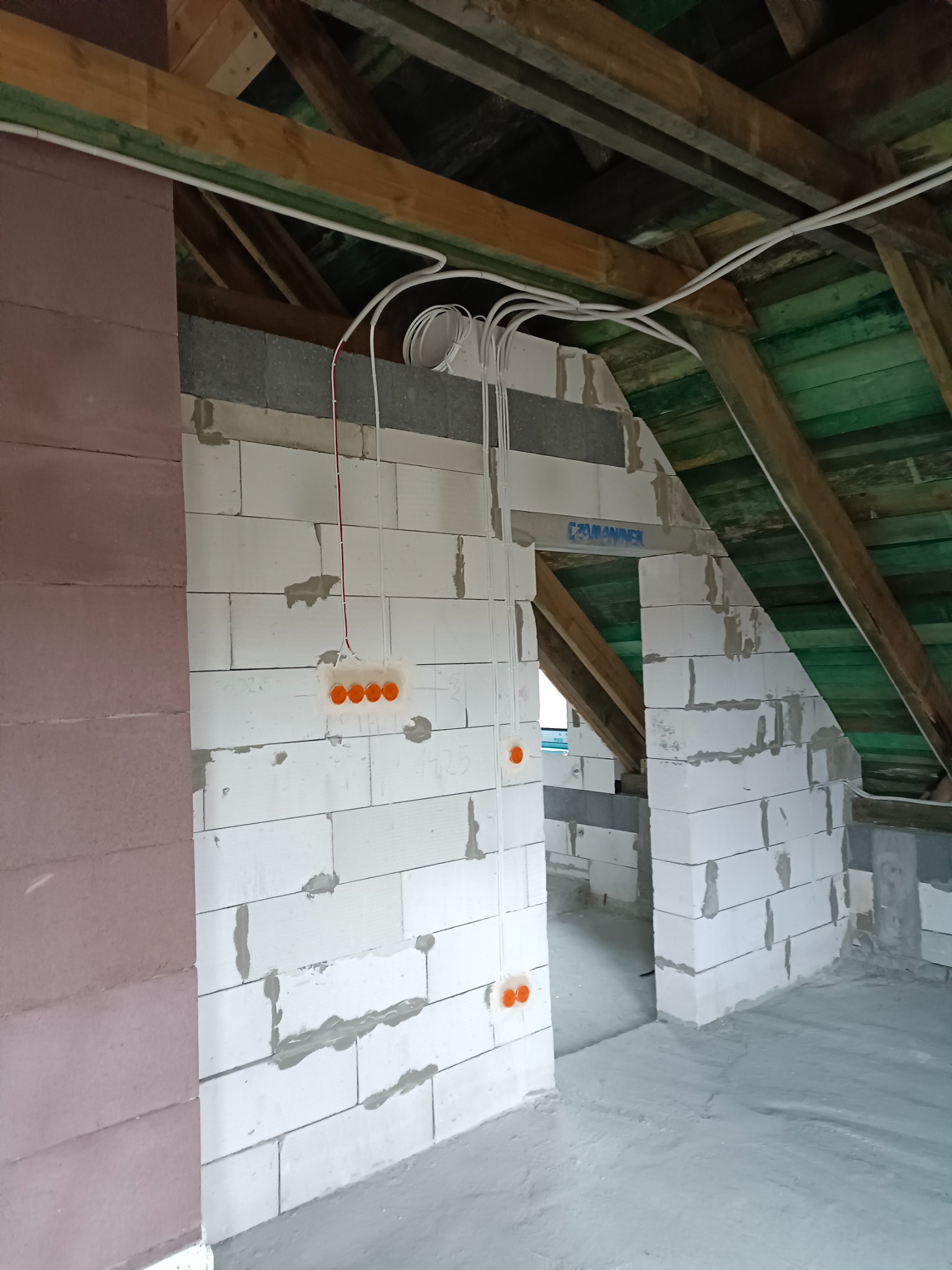 Wnętrze poddasza w trakcie prac wykończeniowych: widoczne białe ściany z betonu komórkowego z poprowadzonymi przewodami elektrycznymi i pomarańczowymi puszkami instalacyjnymi, drewniana konstrukcja...