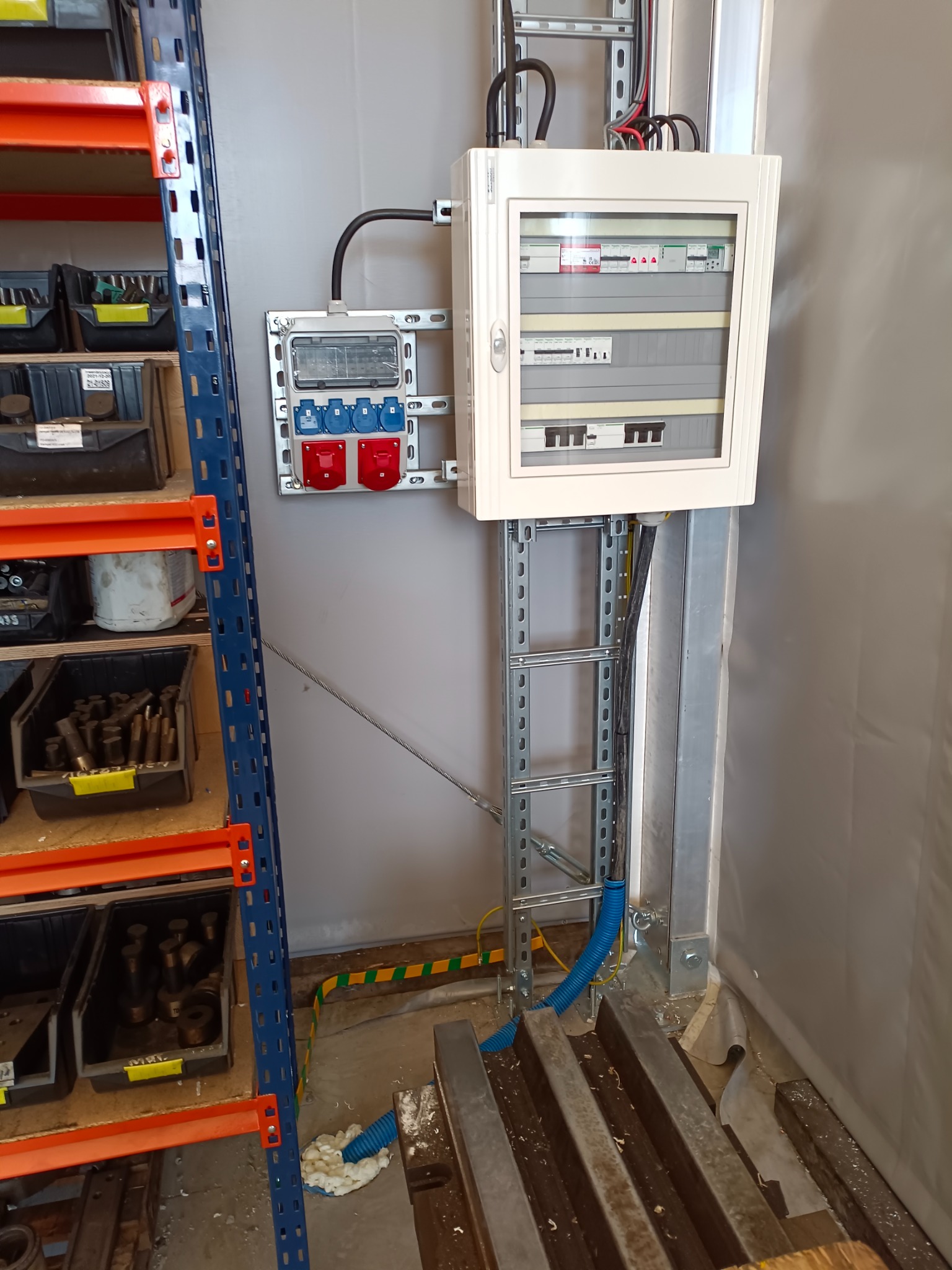 Zamontowana skrzynka elektryczna z bezpiecznikami i gniazdami siłowymi na metalowej konstrukcji w pomieszczeniu warsztatowym, obok widoczny regał z narzędziami.