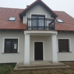 Nowo otynkowany, dwukondygnacyjny dom jednorodzinny w Chodzieży z balkonem i czerwoną dachówką, okna z brązowymi ramami i roletami zewnętrznymi.
