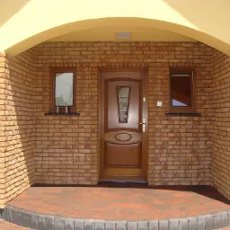 Wejście do domu z brązowymi drzwiami, obłożone cegłą klinkierową i małym oknem po obu stronach, pod łukowatym zadaszeniem.