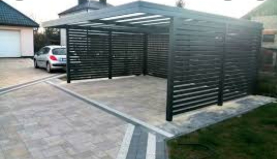 Nowoczesna, grafitowa konstrukcja z poziomych listew, tworząca zadaszone miejsce parkingowe na posesji z kostki brukowej, obok domu z białym garażem i zaparkowanym srebrnym samochodem.