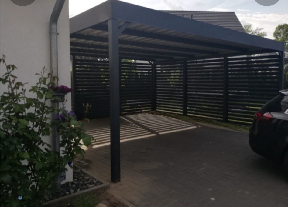 Nowoczesna wiata garażowa z czarnymi, poziomymi panelami, rzucająca cienie na brukowaną nawierzchnię, z fragmentem czarnego samochodu w tle i roślinnością z boku.