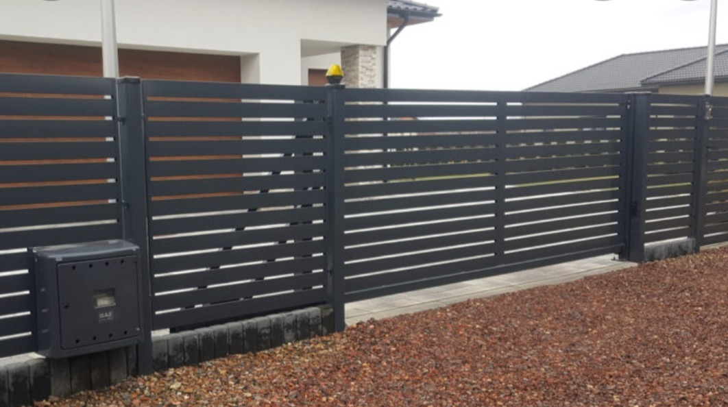 Nowoczesne, poziome, szare przęsła ogrodzeniowe z bramą przesuwną i skrzynką gazową na tle domu z brązowymi elementami elewacji, na podłożu z kory.