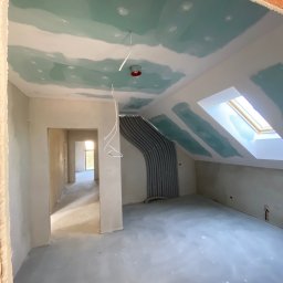 KUB - HOUSE Jakub Piechowicz - Poddasze w trakcie prac wykończeniowych: ściany i sufit pokryte wstępną warstwą gładzi, widoczne rury instalacji, okno dachowe i otwór drzwiowy do kolejnego pomieszczenia.