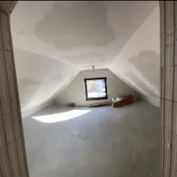 Poddasze w trakcie prac wykończeniowych, widoczne ślady po łataniu ubytków na ścianach i suficie, surowa posadzka, okno dachowe i materiały budowlane w tle.