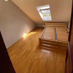 KUB - HOUSE Jakub Piechowicz - Poddasze z drewnianą podłogą i niepomalowanymi szarymi ścianami, widok na drewniane łóżko bez materaca i otwarte okno dachowe wpadające do wnętrza promieniami słońca.